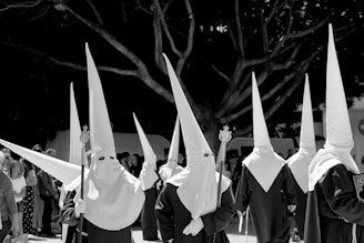 A group of individuals dressed in traditional robes and pointed hoods, participating in a procession. They are holding staffs and the background features a large tree and a crowd of onlookers.