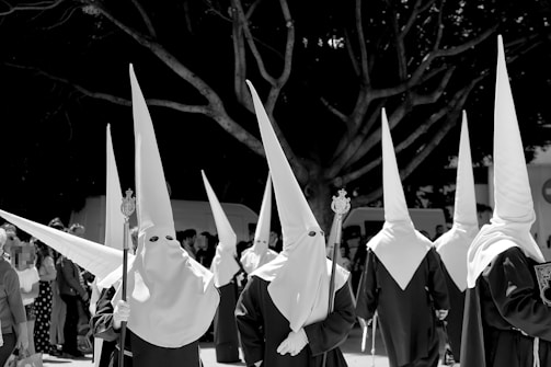 A group of individuals dressed in traditional robes and pointed hoods, participating in a procession. They are holding staffs and the background features a large tree and a crowd of onlookers.