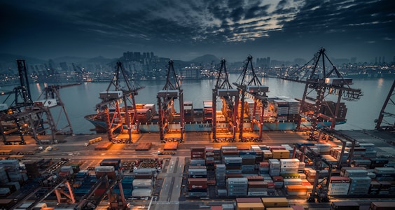 A bustling shipping port is illuminated in the evening, with large cranes towering over numerous shipping containers. The sky is overcast with dramatic clouds, and the lights from the city and port reflect off the water. The atmosphere conveys a sense of industry and activity.