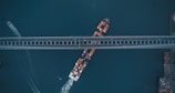 An aerial view depicts a container ship navigating a waterway beneath a large bridge. The ship is laden with multicolored containers, and a small vessel is visible leaving a trail nearby. The bridge is parallel to the bottom edge of the image, and a section of the coastline can be seen on the right.