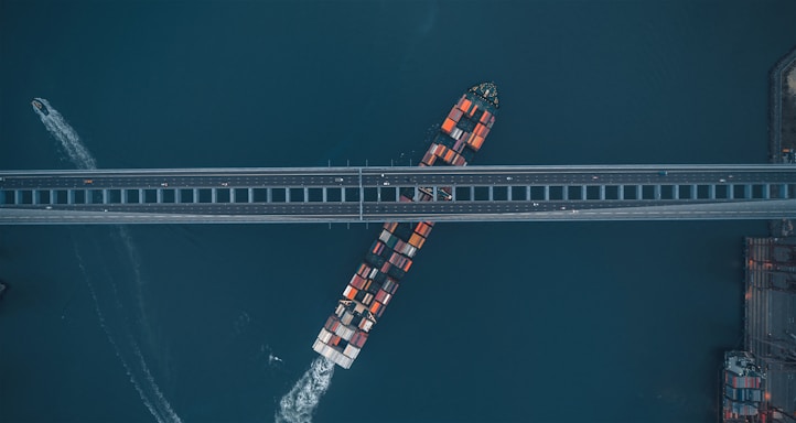 An aerial view depicts a container ship navigating a waterway beneath a large bridge. The ship is laden with multicolored containers, and a small vessel is visible leaving a trail nearby. The bridge is parallel to the bottom edge of the image, and a section of the coastline can be seen on the right.