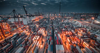 A busy shipping port with stacked containers and cranes under a dark cloudy sky