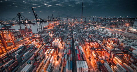 A busy shipping port with stacked containers and cranes under a dark cloudy sky