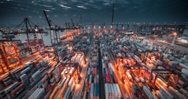 A bustling shipping port at twilight, filled with numerous colorful cargo containers neatly arranged in rows. Shipping cranes tower over the scene, and bright artificial lights create a stark contrast with the dark, cloudy sky. A bridge is visible in the background, adding depth to the composition.