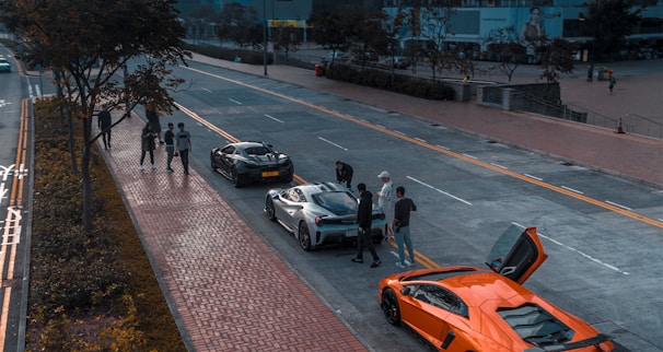 A group of people are gathered around a black sports car in the middle of a street. An orange sports car is parked nearby with its door open. Several other people are walking on the sidewalk lined with trees. The background shows buildings and an open area, with muted urban surroundings.