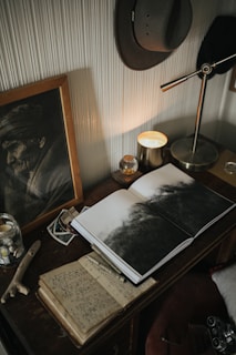 An atmospheric interior setting features a wooden desk adorned with an open book displaying a monochrome image, a handwritten journal, and a framed portrait of an elderly man. A candle emits a warm glow next to a metallic desk lamp, and a hat hangs on the textured wall above the scene.