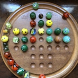 A wooden board with a circular pattern of holes, holding colorful marbles. The marbles are of various colors including green, yellow, blue, and red with some having patterns like dots or wavy lines.