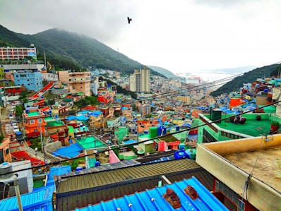 A colorful hillside neighborhood filled with tightly packed houses, painted in various bright colors like blue, green, red, and orange. The area is densely populated, with a mix of multi-story buildings and smaller homes. In the background, there are green hills shrouded in mist and a view of a port with a few ships docked. A bird is flying in the sky above the scene.