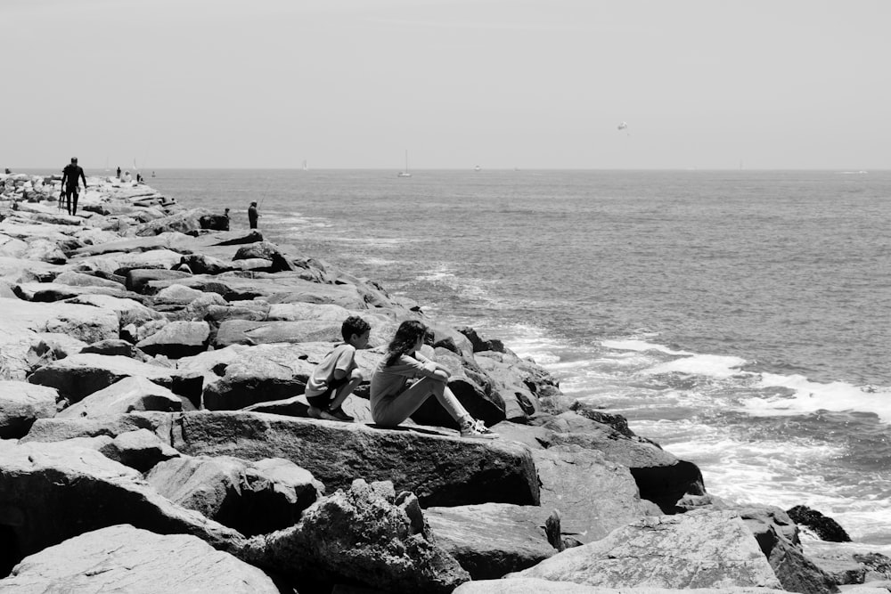 grayscale photo of man and woman sitting on rock near sea