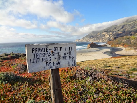 A coastline featuring a scenic view of the ocean bordered by cliffs and a beach. The foreground displays vibrant coastal vegetation and a weathered wooden sign warning against trespassing. The sky is partly cloudy, adding depth to the picturesque landscape.