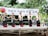 Five women in traditional attire are performing a dance on an outdoor stage. They are wearing black dresses with red accents, holding small objects, and surrounded by trees. A banner above reads 'Festival of Lotus & Asian Cultures' in English and Korean.