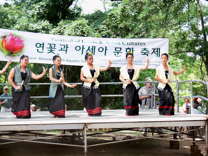 Five women in traditional attire are performing a dance on an outdoor stage. They are wearing black dresses with red accents, holding small objects, and surrounded by trees. A banner above reads 'Festival of Lotus & Asian Cultures' in English and Korean.
