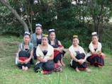 Six young women in traditional attire are sitting on grass in an outdoor setting with trees in the background. They are wearing outfits with black skirts, red accents, yellow and white shawls, and floral headpieces. Their expressions are calm and they are posed for a group photograph.