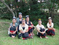 Six young women in traditional attire are sitting on grass in an outdoor setting with trees in the background. They are wearing outfits with black skirts, red accents, yellow and white shawls, and floral headpieces. Their expressions are calm and they are posed for a group photograph.