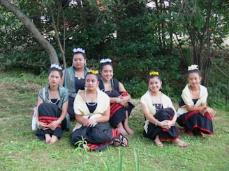 Six young women in traditional attire are sitting on grass in an outdoor setting with trees in the background. They are wearing outfits with black skirts, red accents, yellow and white shawls, and floral headpieces. Their expressions are calm and they are posed for a group photograph.