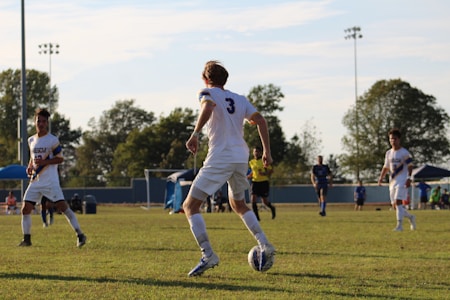 A soccer game taking place on a grassy field with several players in motion. The focus is on a player wearing a white jersey with the number 3, preparing to kick a soccer ball. Other players, some wearing white and others dark blue, are visible in the background along with a referee and spectators on the sidelines. Trees and goal posts are also visible in the background under a partly cloudy sky.