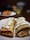 Two rolled crepes filled with cream are placed on a white plate. The crepes appear golden brown, indicating they are perfectly cooked, and are topped with a generous amount of whipped cream or a similar topping. In the background, there is an indistinct, blurred out area with another plate that holds a few out-of-focus items.