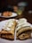 Two rolled crepes filled with cream are placed on a white plate. The crepes appear golden brown, indicating they are perfectly cooked, and are topped with a generous amount of whipped cream or a similar topping. In the background, there is an indistinct, blurred out area with another plate that holds a few out-of-focus items.