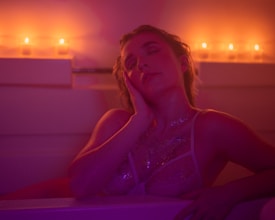A person is relaxing in a bathtub with closed eyes, leaning their head on their hand. The room is dimly lit with warm orange and purple hues, creating a cozy atmosphere. Candles are placed in the background, adding to the serene and intimate ambiance. The person is adorned with body glitter and is wearing delicate lingerie.
