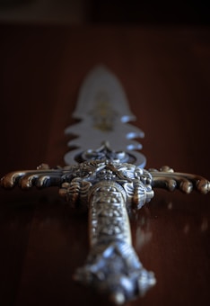 An ornate sword with intricate detailing on the hilt and a shiny, reflective blade is placed on a wooden surface. The design features elaborate patterns and metallic embellishments that give it an antique and ceremonial appearance.