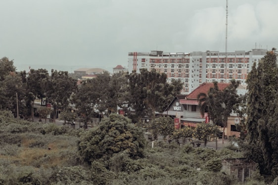An urban landscape features a mix of greenery and modern architecture. There is a row of trees in the foreground, a dense growth of bushes and plants, and several multi-story buildings in the background. One of the buildings has a sign with the word 'VAST', suggesting it may be a business or public space.