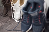 A group of shoes is arranged on a wooden floor. The collection includes three pairs: dark brown boots, white sneakers with white laces, and dark sneakers with red accents.