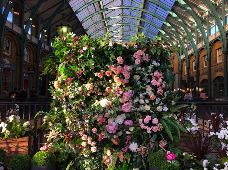 A lush, vertical display of assorted flowers, predominantly pink and white, surrounded by vibrant green foliage. This floral arrangement is set against an elegant arched indoor setting with a glass-paneled ceiling and ornate metal structures, providing a charming contrast between nature and architecture.