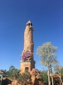 A tall, cylindrical stone tower with a rustic appearance stands against a bright blue sky. The tower features a sign reading 'Islands of Adventure' in bold, colorful letters. Surrounding the base of the tower are lush green trees and foliage, creating a vibrant and inviting atmosphere.
