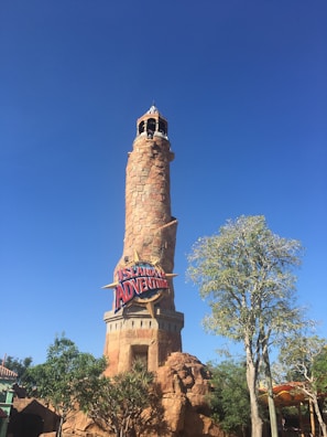 A tall, cylindrical stone tower with a rustic appearance stands against a bright blue sky. The tower features a sign reading 'Islands of Adventure' in bold, colorful letters. Surrounding the base of the tower are lush green trees and foliage, creating a vibrant and inviting atmosphere.