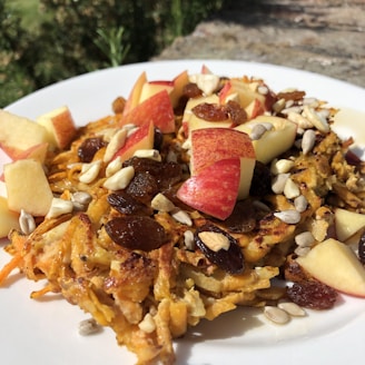 A white plate holds a portion of grated vegetable fritters, topped with diced red apples, raisins, and a mix of sunflower seeds and chopped nuts. The dish sits outdoors with greenery in the background, catching sunlight which highlights the textures and colors of the ingredients.