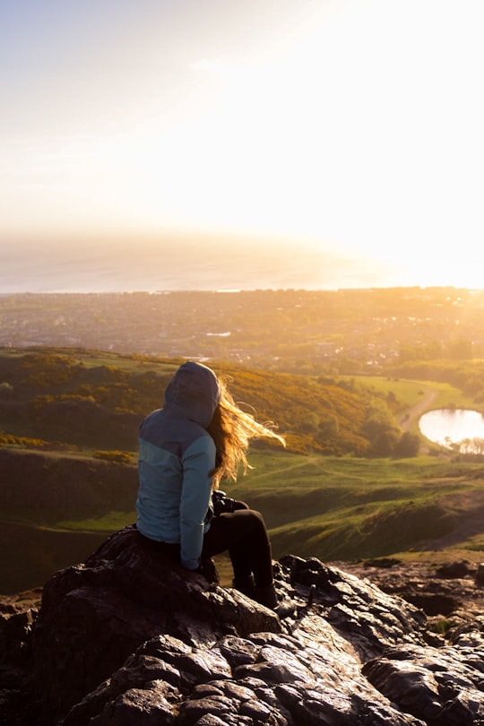 woman in gray hoodie sitting on rock during daytime in Arthur's Seat United Kingdom