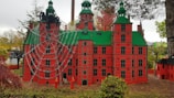 A detailed model of a red building with green roofs, adorned with large spider figures and a web across the facade. The background features trees and grassy areas, contributing to a whimsical outdoor setting.