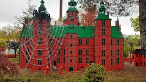 A detailed model of a red building with green roofs, adorned with large spider figures and a web across the facade. The background features trees and grassy areas, contributing to a whimsical outdoor setting.