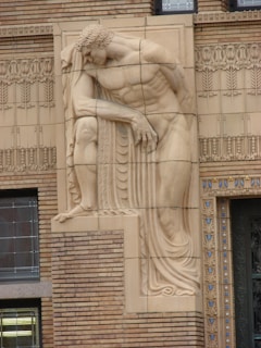 A detailed bas-relief sculpture of a muscular, reclining male figure is integrated into the intricate architectural design of a building facade. The sculpture showcases fine details in the man's hair and drapery, blending classical elements with the linear patterns of the surrounding brickwork and ornate decoration.