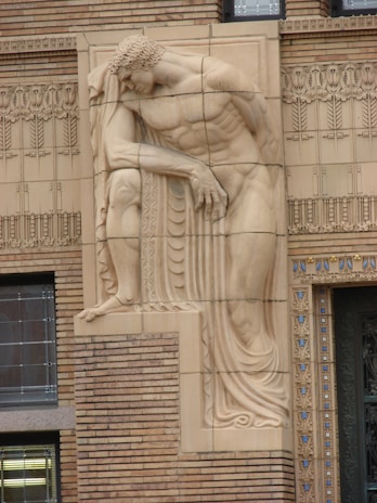 A detailed bas-relief sculpture of a muscular, reclining male figure is integrated into the intricate architectural design of a building facade. The sculpture showcases fine details in the man's hair and drapery, blending classical elements with the linear patterns of the surrounding brickwork and ornate decoration.