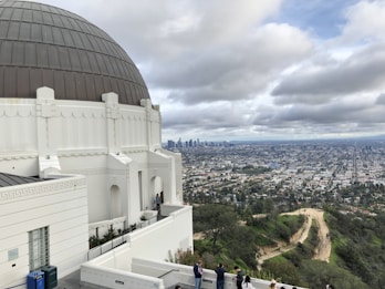 A large white observatory building with a domed roof is situated on a hill, offering a panoramic view of a sprawling cityscape. The skyline includes numerous skyscrapers in the distance. Several people are observing the view from the terrace of the observatory. The sky is overcast with thick clouds.