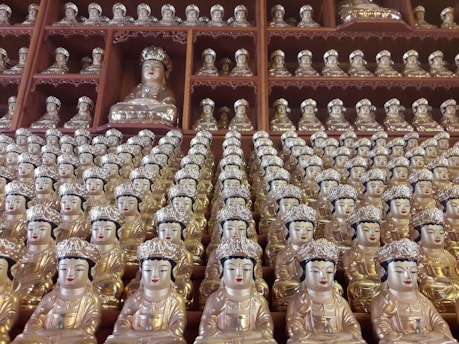 A multitude of small, intricately designed golden and silver statues lined up in rows and columns. Each statue depicts a serene, meditative figure with a crown-like headpiece and detailed facial features. The statues are arranged in a wooden shelf structure, creating an impression of abundance and unity.