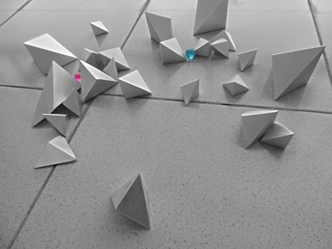 Geometric shapes crafted from paper in the form of pyramids scattered across a tiled floor. The scene includes two colorful gems, one turquoise and one pink, nestled among the shapes.