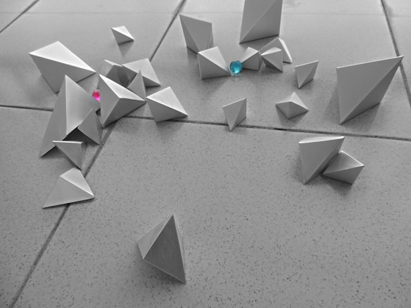 Geometric shapes crafted from paper in the form of pyramids scattered across a tiled floor. The scene includes two colorful gems, one turquoise and one pink, nestled among the shapes.