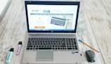 A laptop is open on a wooden table displaying a website, with a mouse and a pen positioned to its right side. Two highlighters, one blue and one purple, are to the left of the laptop.
