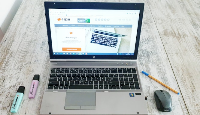 A laptop is open on a wooden table displaying a website, with a mouse and a pen positioned to its right side. Two highlighters, one blue and one purple, are to the left of the laptop.