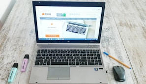 A laptop is open on a wooden table displaying a website, with a mouse and a pen positioned to its right side. Two highlighters, one blue and one purple, are to the left of the laptop.