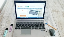 A laptop is open on a wooden table displaying a website, with a mouse and a pen positioned to its right side. Two highlighters, one blue and one purple, are to the left of the laptop.