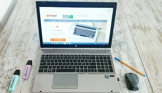 A laptop is open on a wooden table displaying a website, with a mouse and a pen positioned to its right side. Two highlighters, one blue and one purple, are to the left of the laptop.