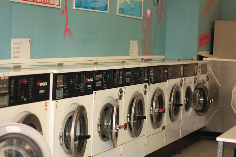 A row of industrial washing machines is lined up against a teal-colored wall, each with digital displays and numbered labels. Above the machines, there are informational posters and light wall decorations.