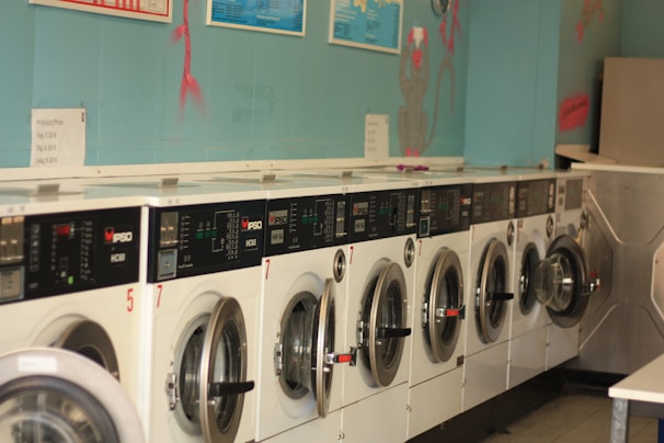 A row of industrial washing machines is lined up against a teal-colored wall, each with digital displays and numbered labels. Above the machines, there are informational posters and light wall decorations.