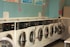 A row of industrial washing machines is lined up against a teal-colored wall, each with digital displays and numbered labels. Above the machines, there are informational posters and light wall decorations.