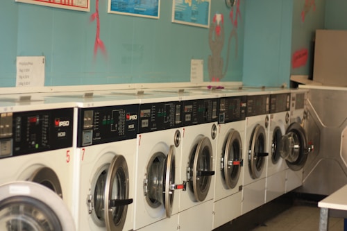 A row of industrial washing machines is lined up against a teal-colored wall, each with digital displays and numbered labels. Above the machines, there are informational posters and light wall decorations.
