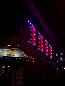 A building exterior at night featuring bright red and blue illuminated lights in a rectangular pattern along the upper wall. Below, a sign with the partial word ‘Roof’ and the phrase ‘Music Joint’ is visible, lit by two overhead lamps. Multi-colored string lights are draped across the awning, enhancing the festive atmosphere.
