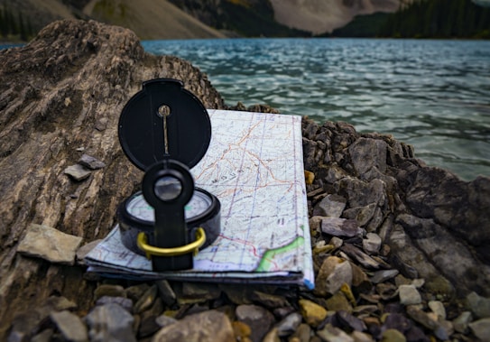 A black compass is placed on top of a detailed map, resting on rocky terrain by the edge of a lake. The lake has clear blue water, and mountainous landscape with forests can be seen in the background.
